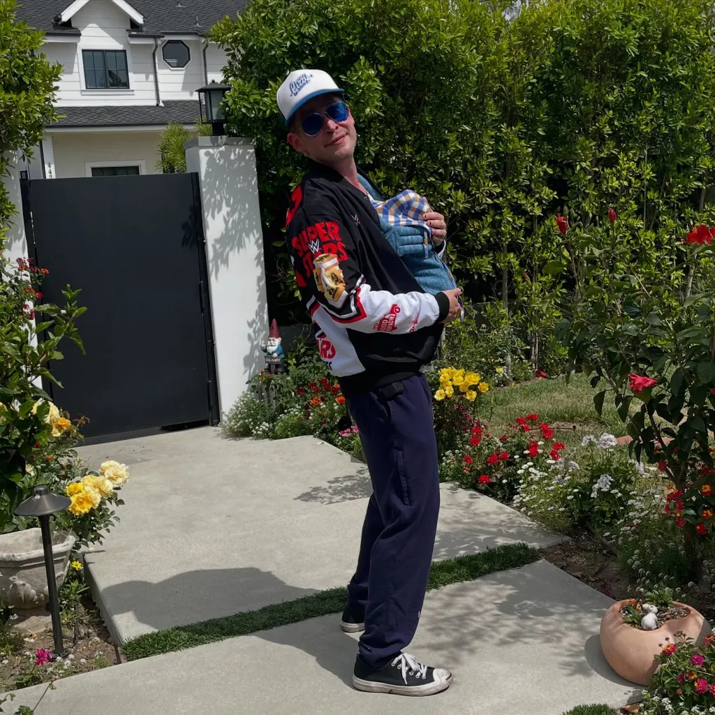 Macaulay Culkin holding a baby in a carrier in a garden.