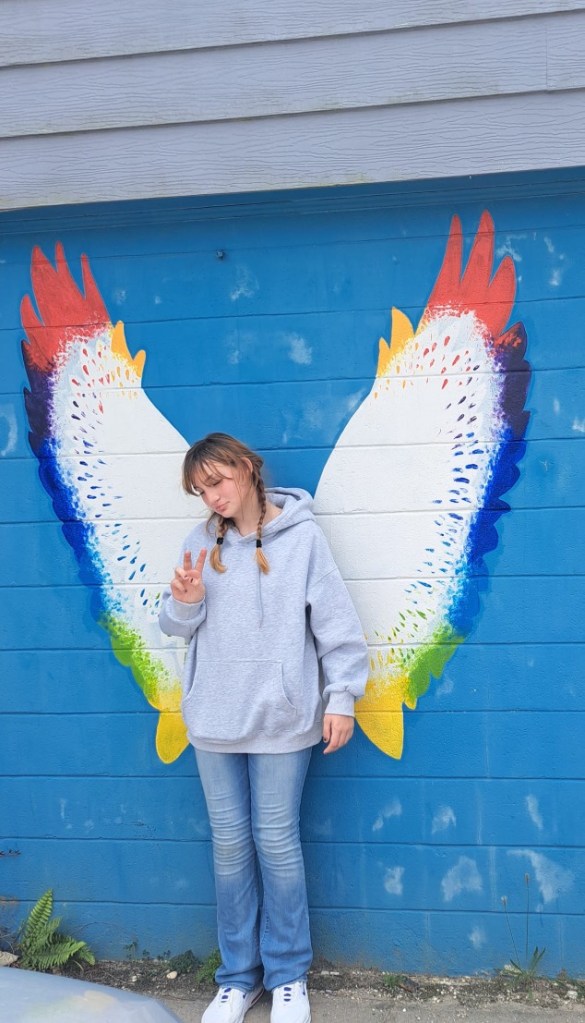 Danika Troy posing in front of a blue wall with rainbow-colored wings.