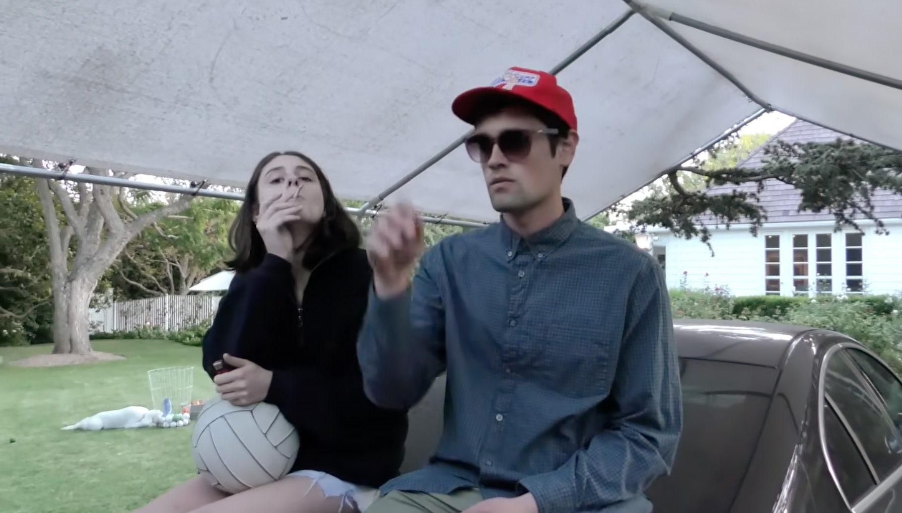 Nick Reiner and a woman sitting on the back of a car under an awning.