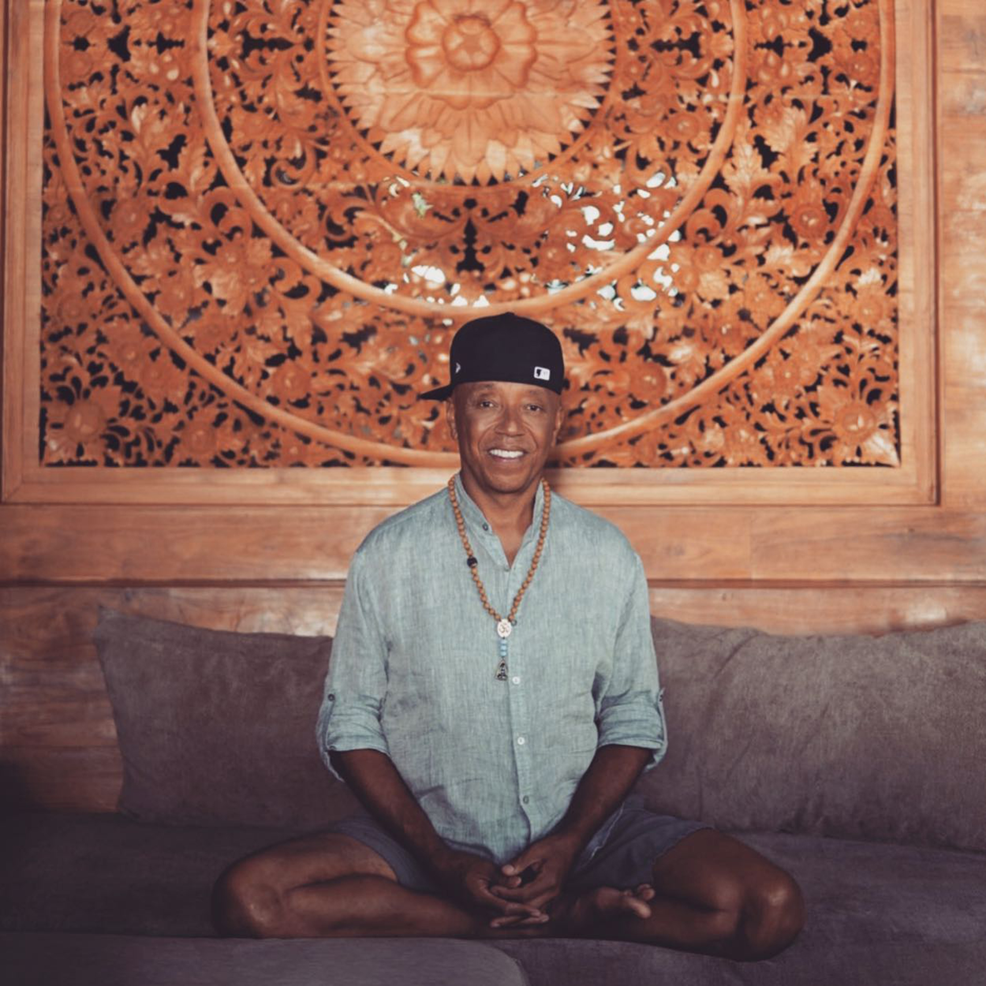 Russell Simmons smiling in a light blue shirt and black baseball cap, sitting cross-legged on a couch in front of an ornate wooden wall carving.
