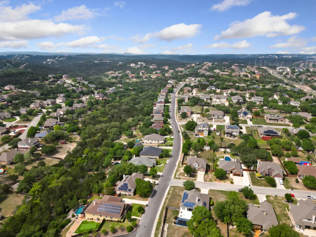 Aerial view of a sprawling residential neighborhood with houses and winding roads nestled among green trees under a partly cloudy sky.