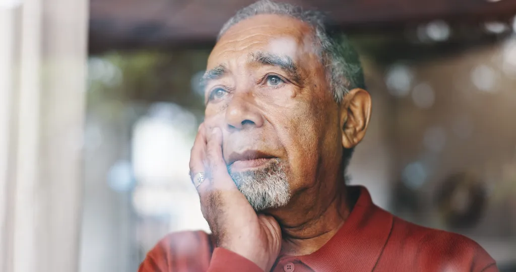 A senior man in a retirement home, looking out a window with a sad expression.