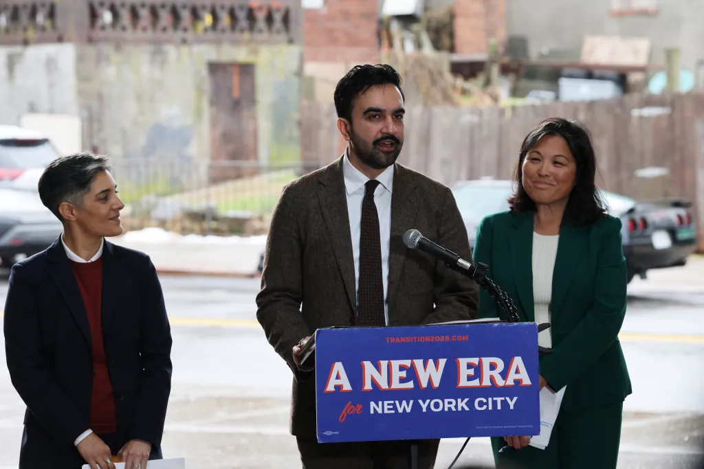New York Mayor-elect Zohran Mamdani speaks at a podium with Leila Bozorg and Julie Su standing beside him.