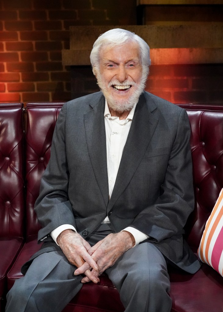 Dick Van Dyke, with white hair and beard, wearing a suit, sits on a red couch and smiles.