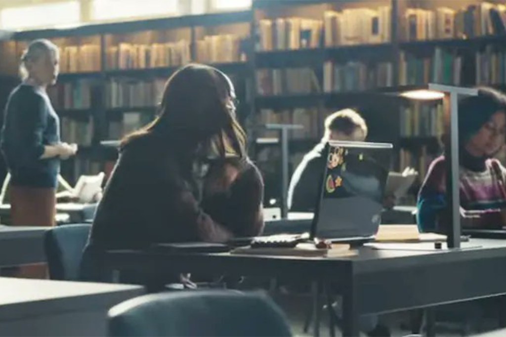 A library scene with a woman's laptop in the foreground displaying a small watermelon sticker.