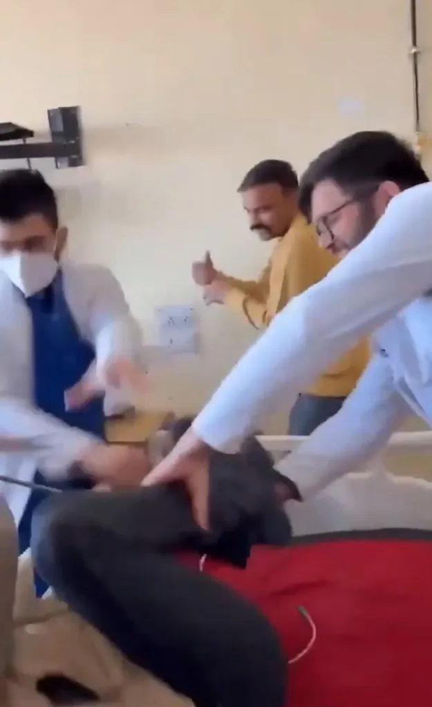 Two doctors beating a patient on a bed, with a third person in the background giving a thumbs-up.