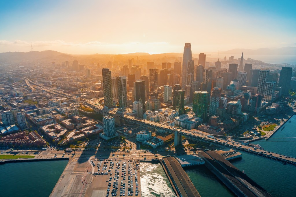 Aerial view of downtown San Francisco with skyscrapers, the Bay Bridge, and surrounding urban landscape at sunrise.