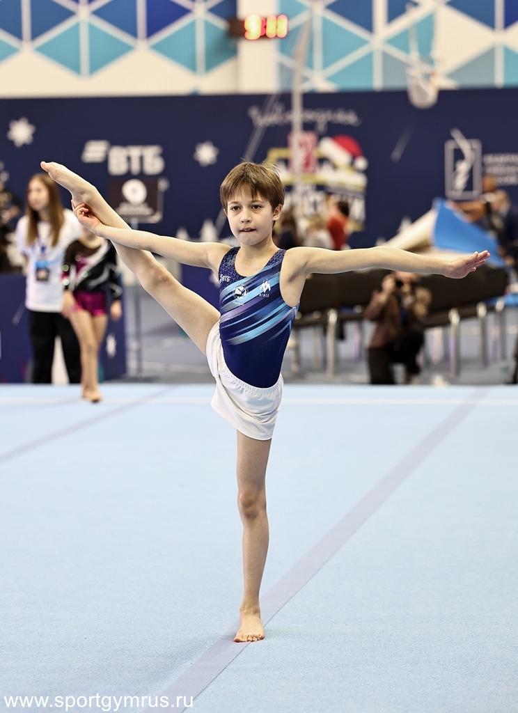 A young male gymnast in a blue leotard and white shorts performs a leg split while balancing on one leg.