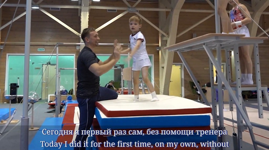 A boy on a mat and a coach clapping his hands in a gym.