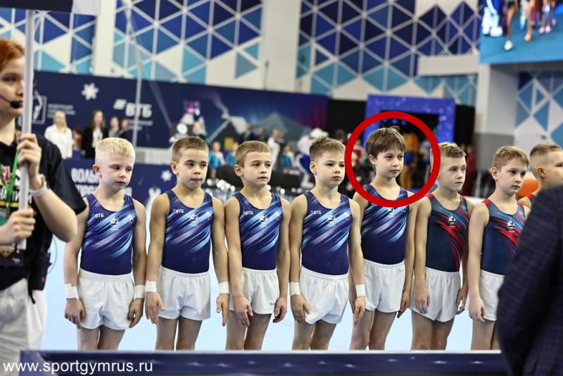 A line of young gymnasts, one circled in red, stands at a competition.