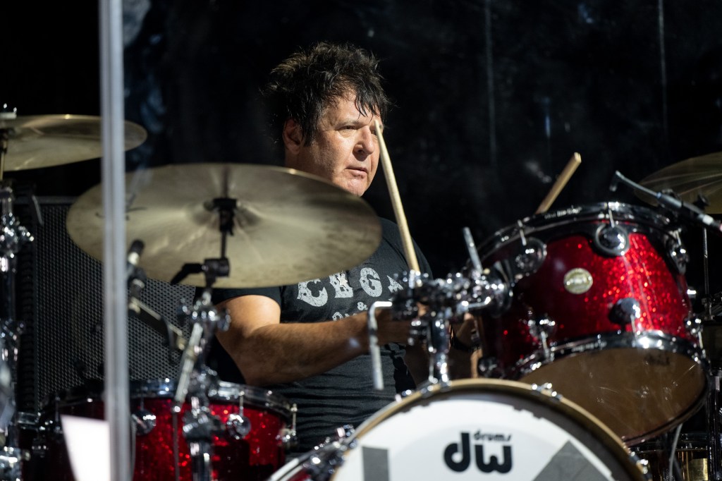 A drummer with shaggy dark hair wearing a black shirt with a white logo behind a red drum kit, focused on playing.