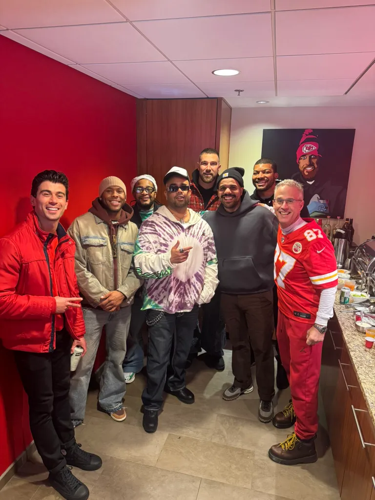 A group of men posing for a photo in a kitchen with a painting of Travis Kelce in the background.