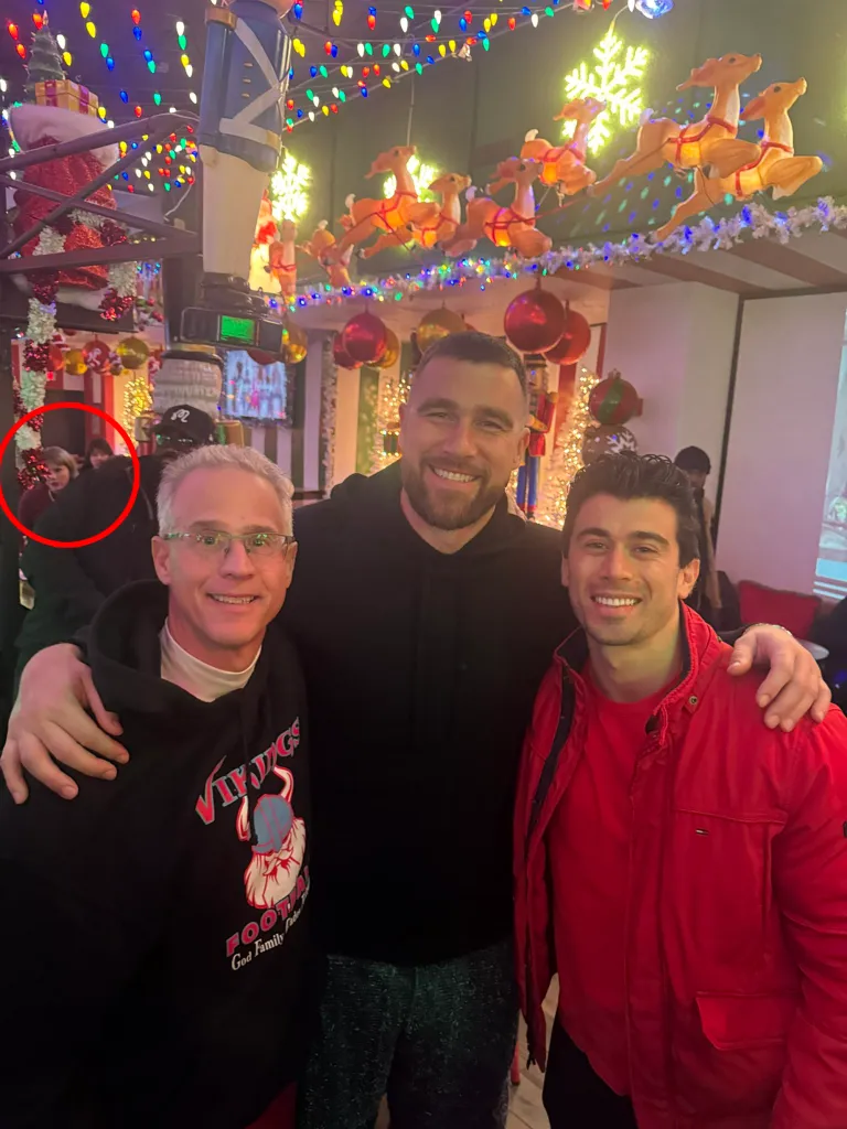 Travis Kelce posing for a photo with two men, with Taylor Swift subtly visible in the background.