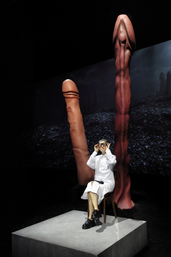 Elke Luyten, dressed in a white lab coat and skirt, sits in a chair between two large phallic sculptures.