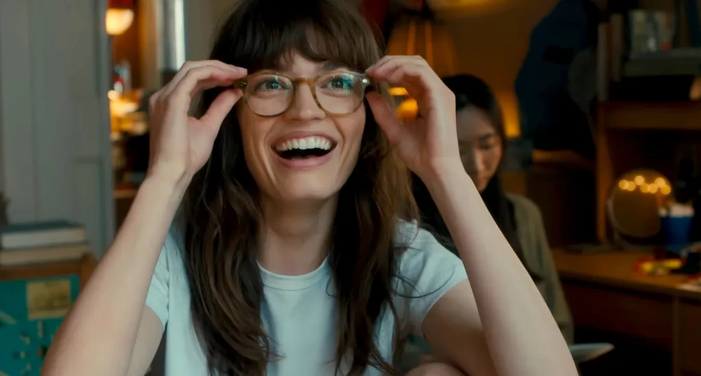 Portrait of Emma Mackey smiling and adjusting her eyeglasses.