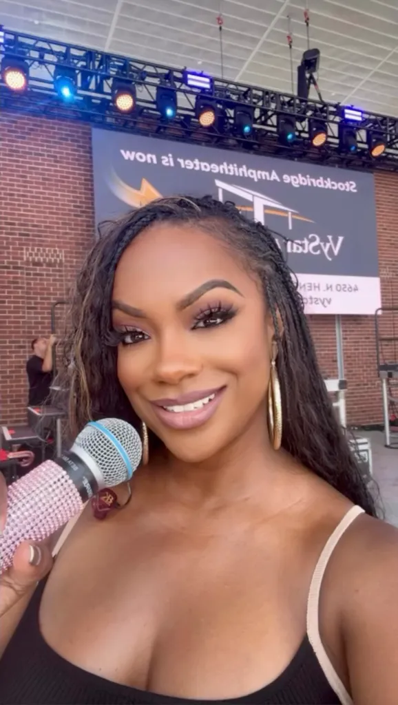 Kandi Burruss holding a pink bejeweled microphone and smiling, with an amphitheater banner in the background.