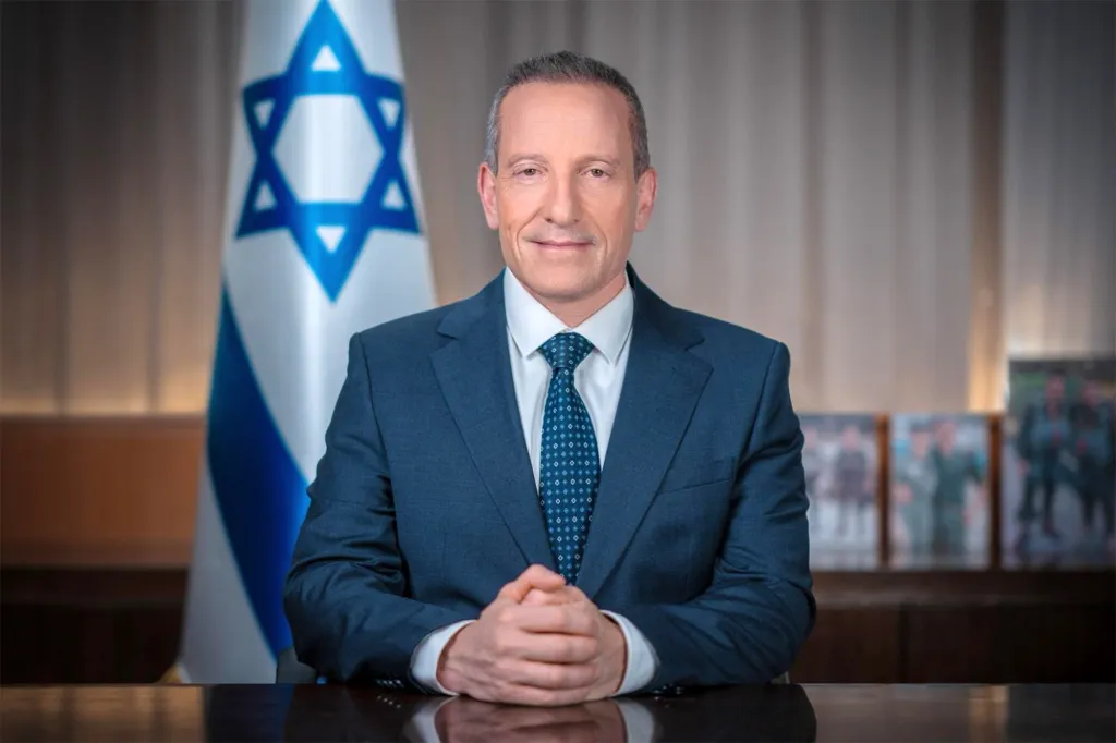 Amir Baram, Director-General of the Israeli Ministry of Defense, in a suit and tie, seated at a desk with the Israeli flag in the background.