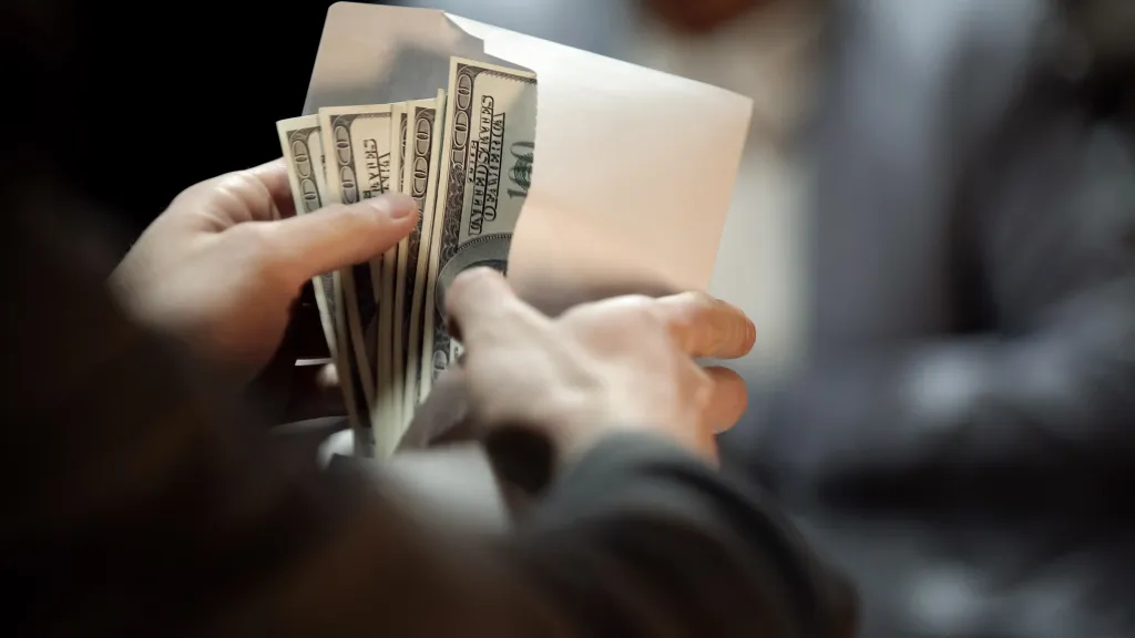 Hands placing US dollar bills into an envelope.