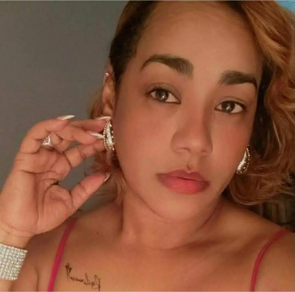 A woman with light brown hair, wearing a pink tank top, a diamond bracelet, and holding an earring.