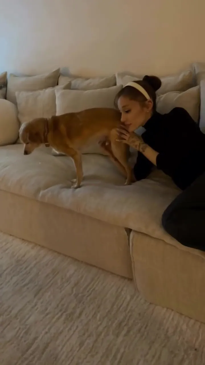 Ariana Grande wearing a headband and black top, affectionately petting a small brown dog on a cream-colored couch.
