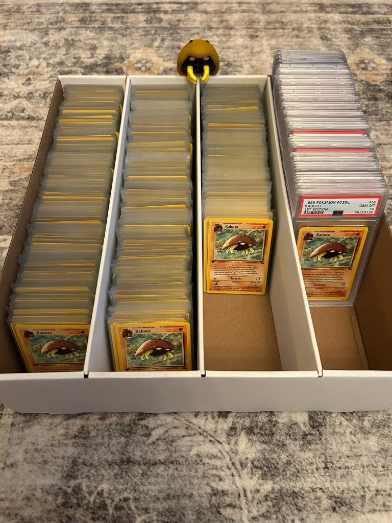 A white storage box filled with many Kabuto Pokemon cards, some loose and some graded, with a small yellow creature peeking over the top.