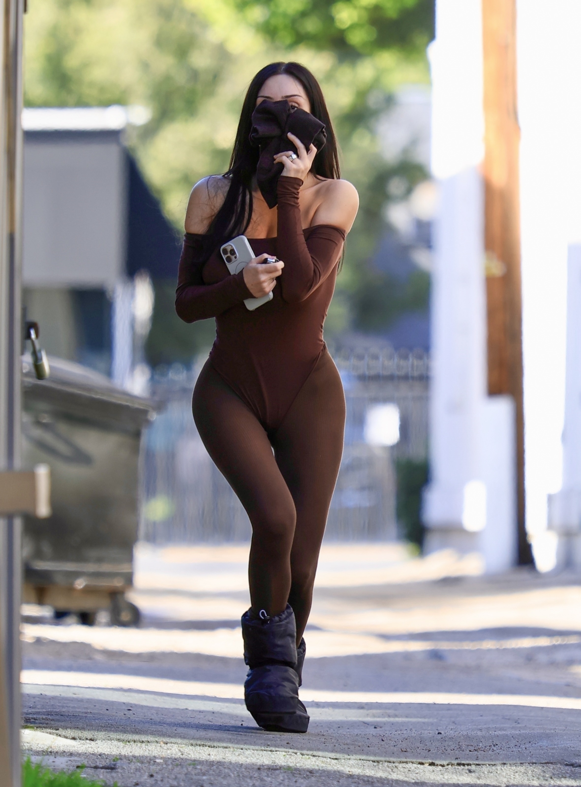 Bianca Censori in a brown bodysuit and moon boots, holding a phone and covering her face with a brown cloth.