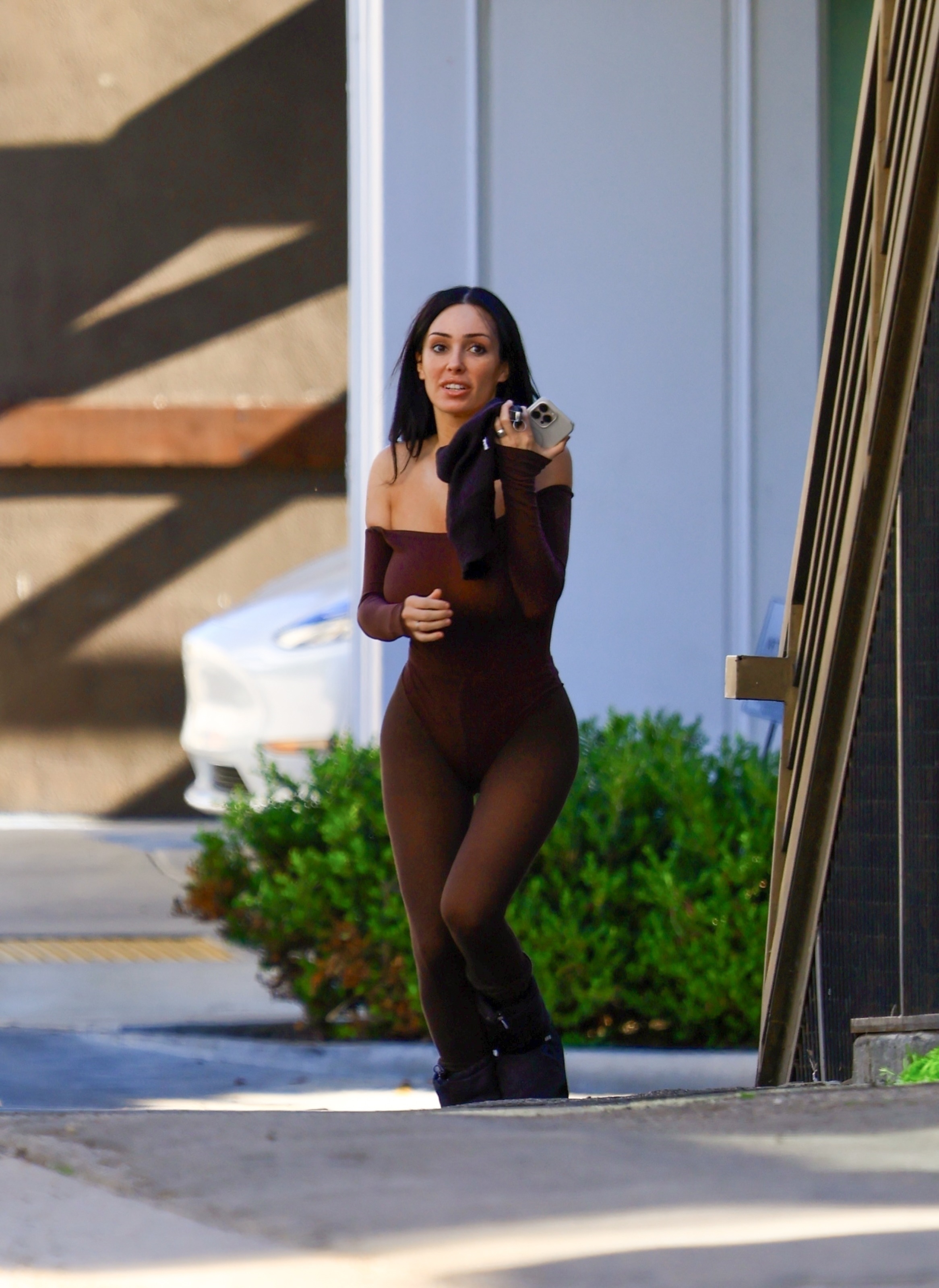 Bianca Censori in a brown bodysuit and Moon Boots holding a phone and shorts.