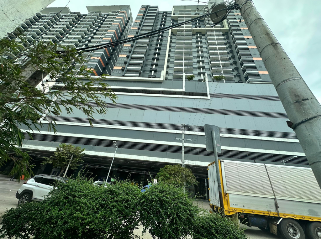 A tall, modern apartment building with multiple floors and balconies, partially obscured by power lines and lush green foliage in the foreground.