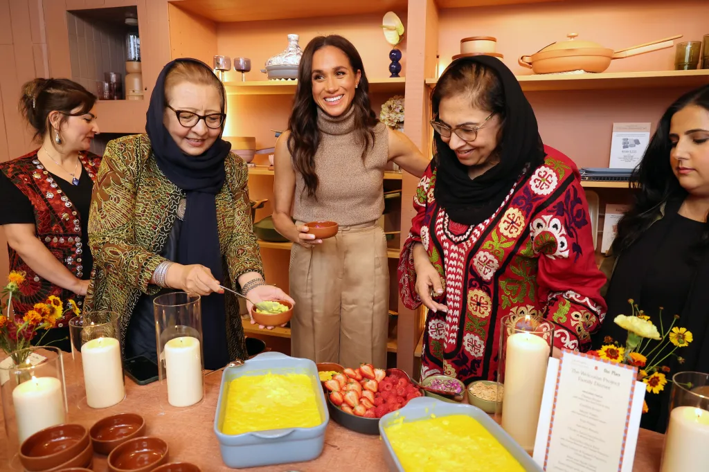 Meghan, Duchess of Sussex, standing with two women and smiling while one woman serves a dish at a holiday dinner.