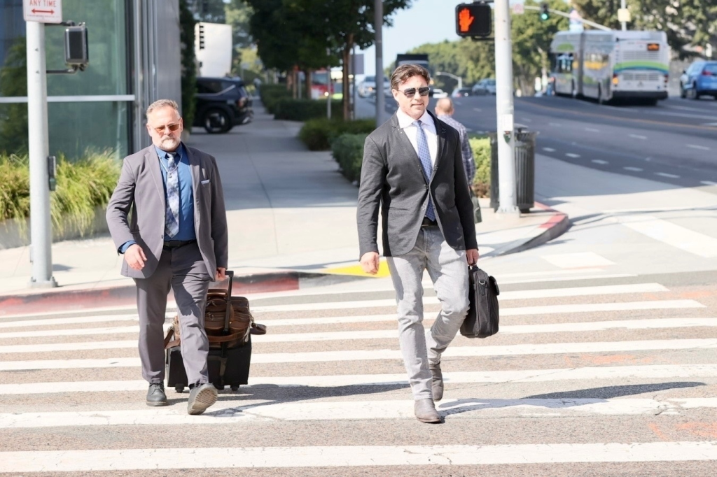 Aaron Phypers in sunglasses and a blazer crossing the street with a briefcase.