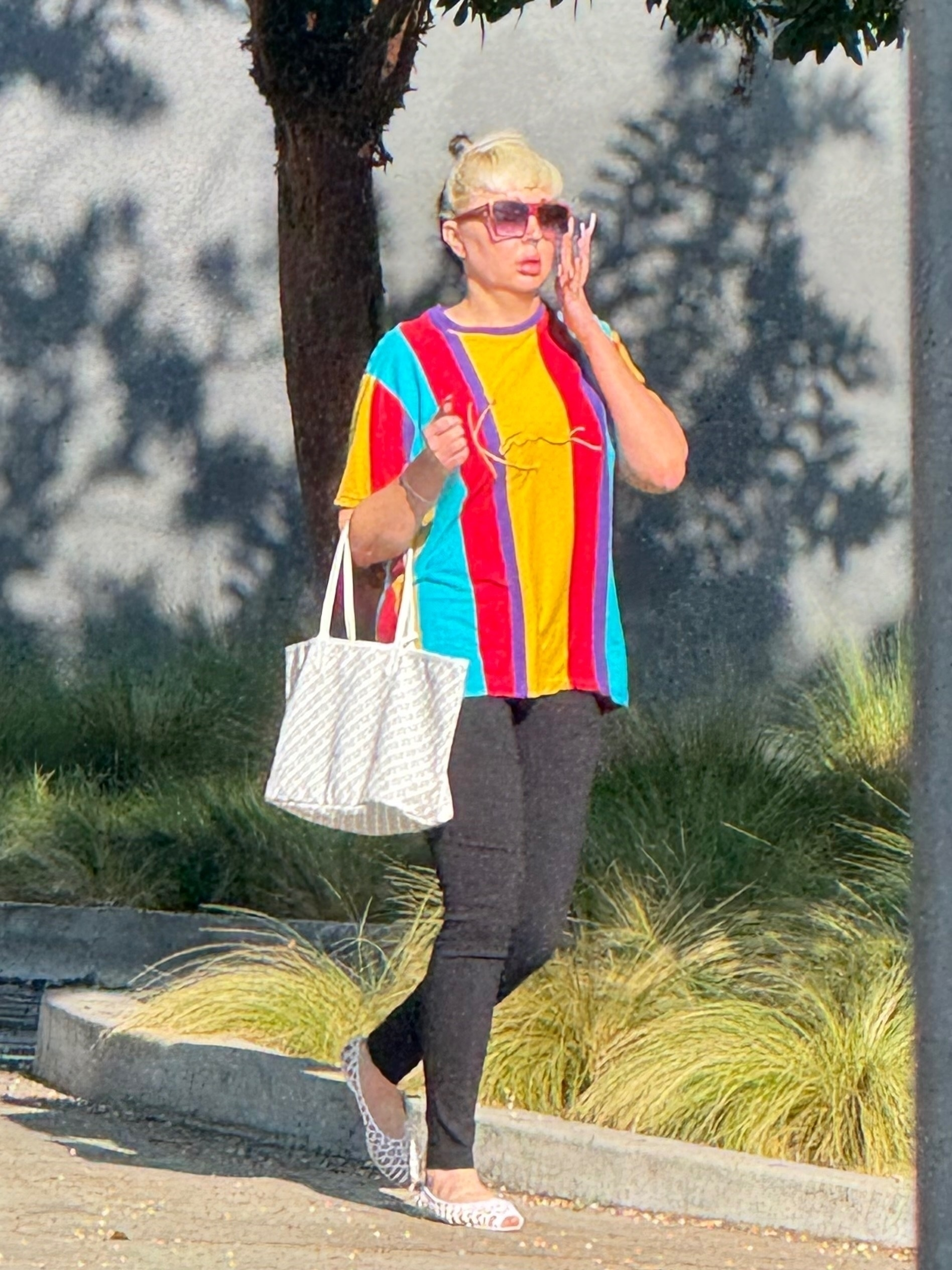 Amanda Bynes in a colorful striped t-shirt and black leggings, carrying a white tote bag and wearing leopard print flats, walking outdoors.