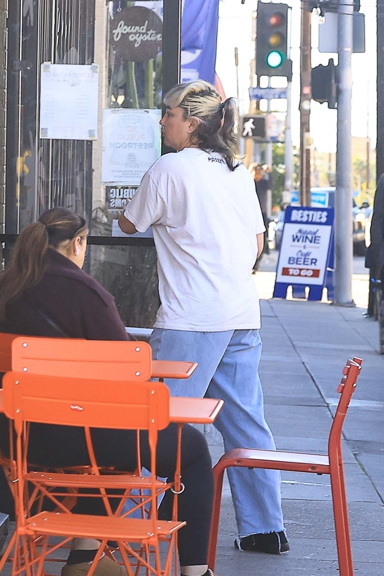 Amanda Bynes in an oversized white shirt and blue jeans, with blonde and dark hair in a ponytail, standing on a sidewalk next to a building.