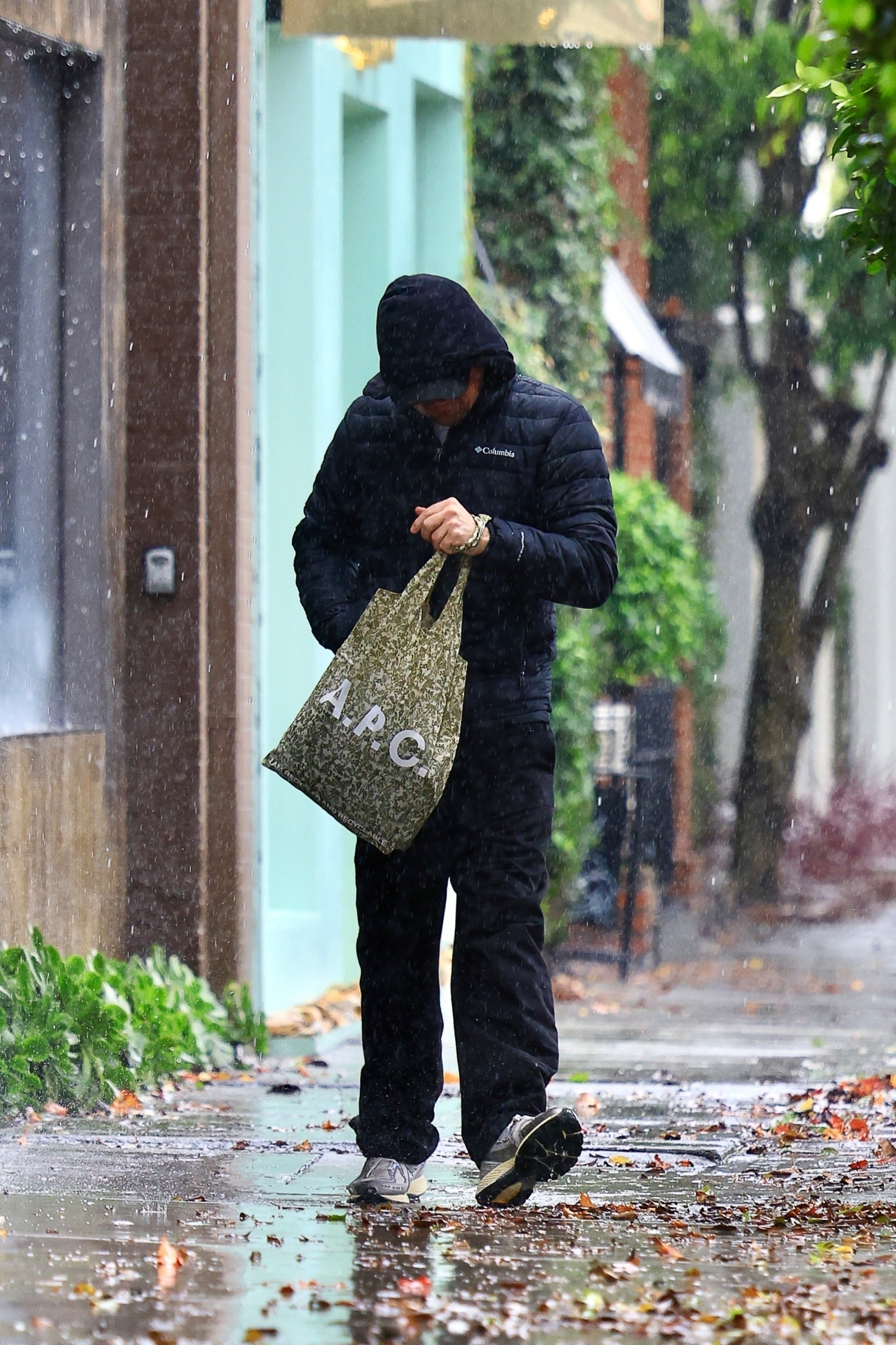 Leonardo DiCaprio walks in the rain, head down and wearing a black hooded jacket, black pants, and a patterned A.P.C. tote bag.