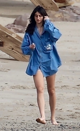 Romy Reiner walking barefoot on Malibu beach, wearing an oversized blue denim shirt.