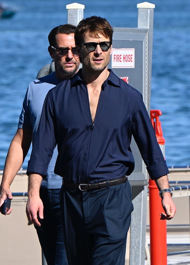 Glen Powell in sunglasses and a navy zipped-up shirt walking by a “Fire Hose” sign.