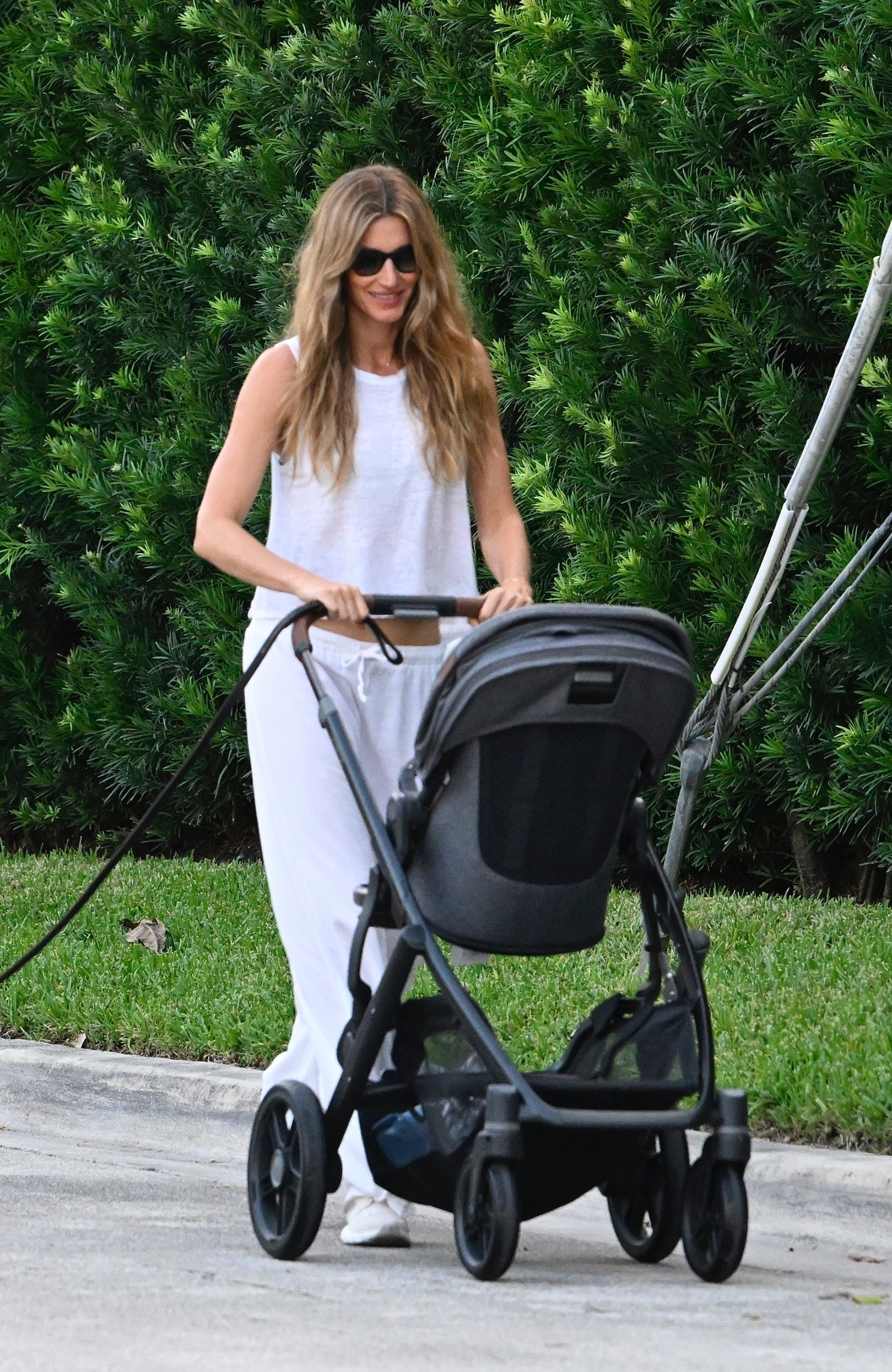 Gisele Bündchen pushing a stroller and holding a dog leash while wearing a white sleeveless top and white pants in Miami.
