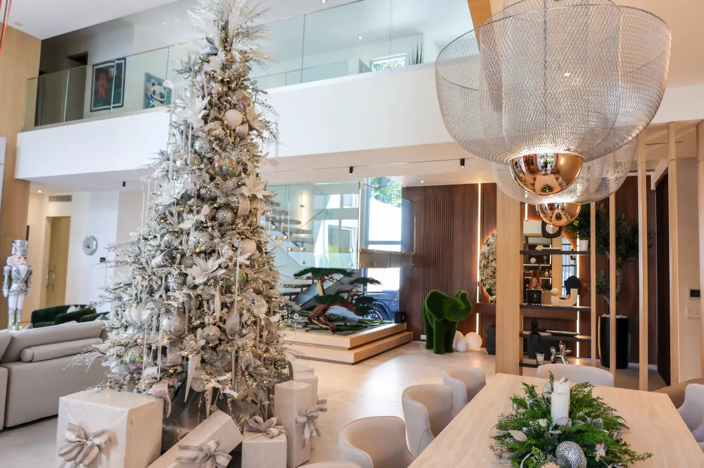 A large, modern living space decorated for Christmas, featuring a tall white and silver Christmas tree with gifts at its base, and a dining table with a festive centerpiece.