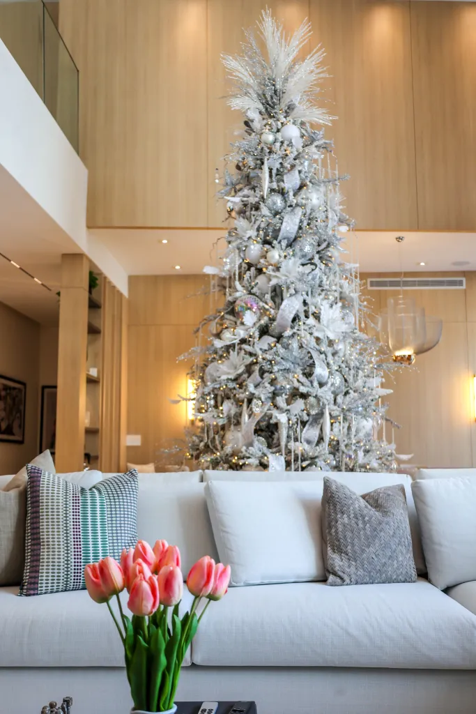 A living room with a white sectional sofa, a vase of pink tulips, and a large Christmas tree decorated with silver ornaments and lights.