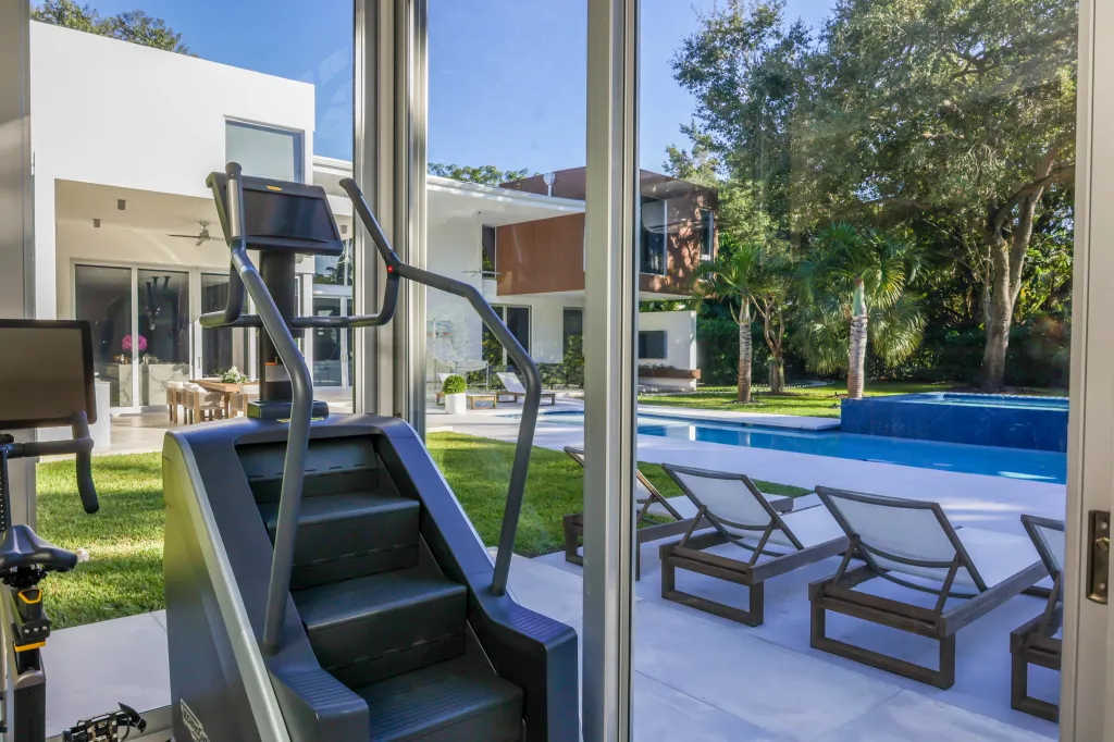 A stair-stepper and exercise bike inside a room with floor-to-ceiling windows looking out onto a backyard with a swimming pool.