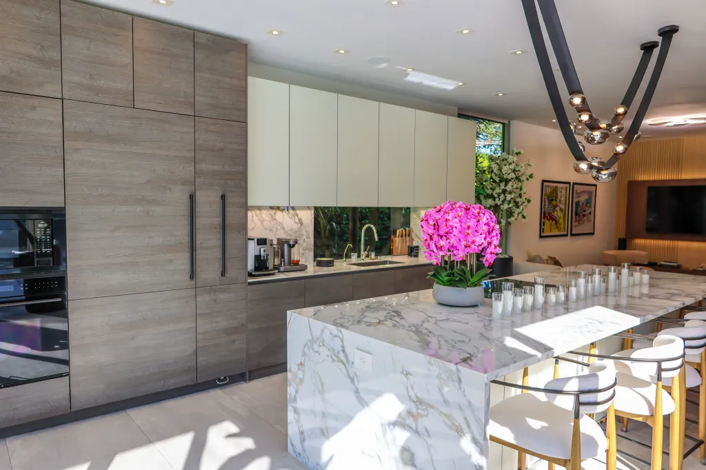 A modern kitchen with light wood-grain cabinets and built-in appliances, a long white marble island with white bar stools and a pink floral centerpiece.