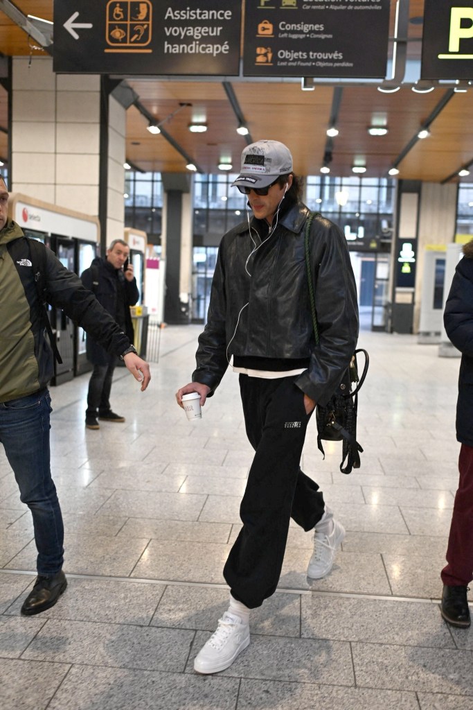 Jacob Elordi departing Gare du Nord in Paris with a coffee and bag, wearing a cap, sunglasses, black leather jacket, and sweatpants.
