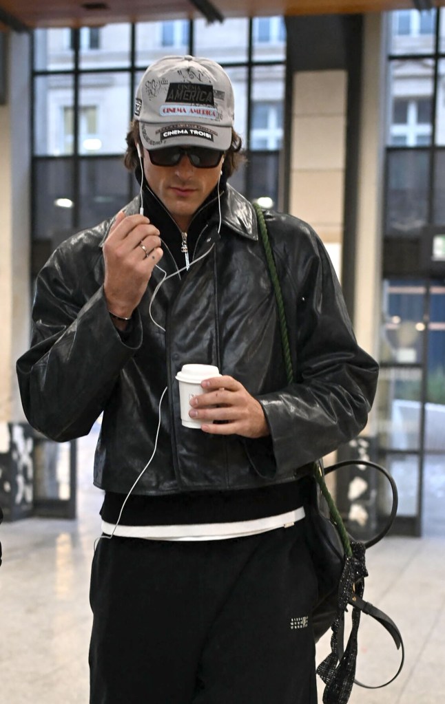 Jacob Elordi holding a coffee and wearing a black leather jacket, gray baseball cap, sunglasses, and earbuds.