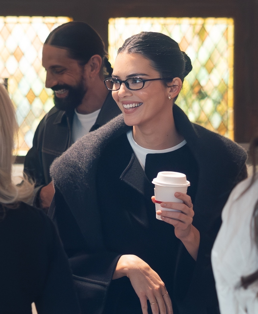 Kendall Jenner smiles, holding a takeaway coffee cup, with perfumer Ben Gorham in the background.