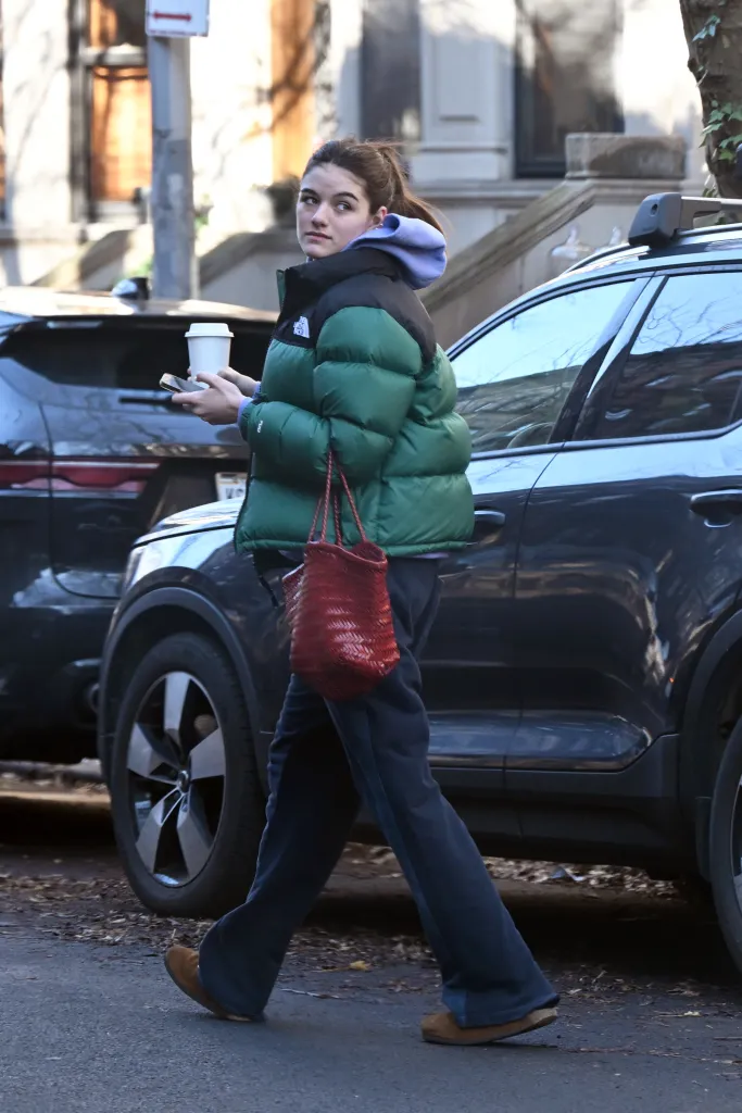 Suri Cruise in a green puffer jacket, black pants, and brown slippers, carrying a red woven bag and a white cup, walking past a parked car.