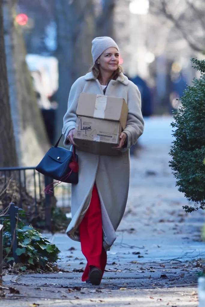 Katie Holmes doing last-minute Christmas shopping while carrying boxes and wearing a beige coat, red trousers, and a beanie hat.