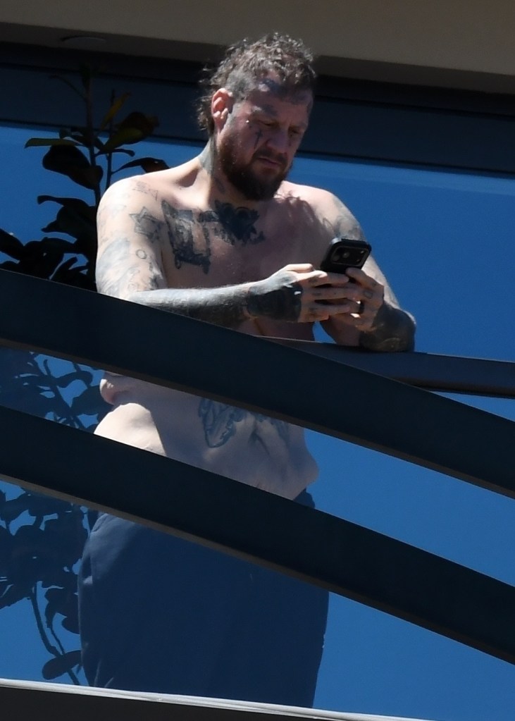 Shirtless Jelly Roll looking at his phone from a balcony.