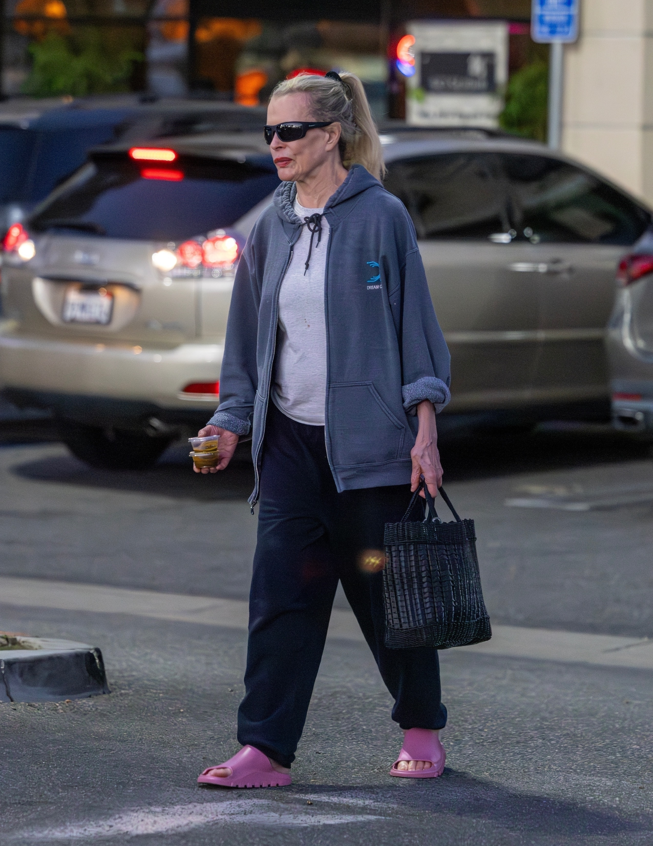 Kim Basinger in a grey hoodie, black sweatpants, pink slides, and black sunglasses, carrying a black bag and small containers of food.