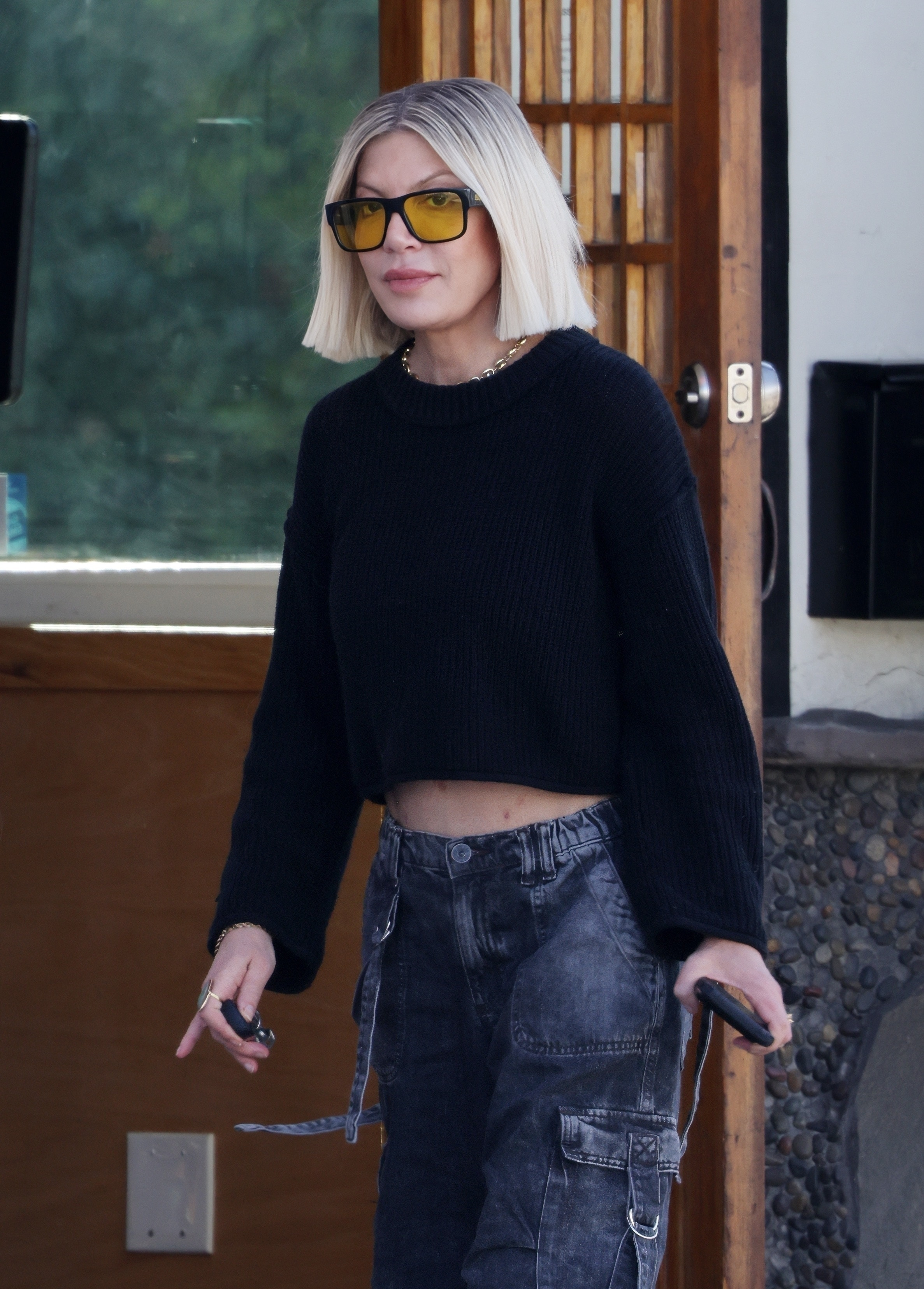 Tori Spelling wearing a black sweater, faded cargo pants, and yellow-tinted sunglasses.