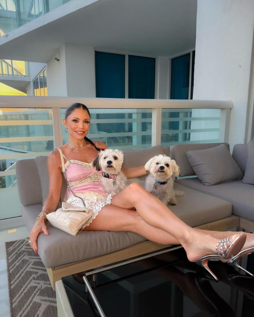 Bethenny Frankel sitting on a couch with two small white dogs on a balcony.