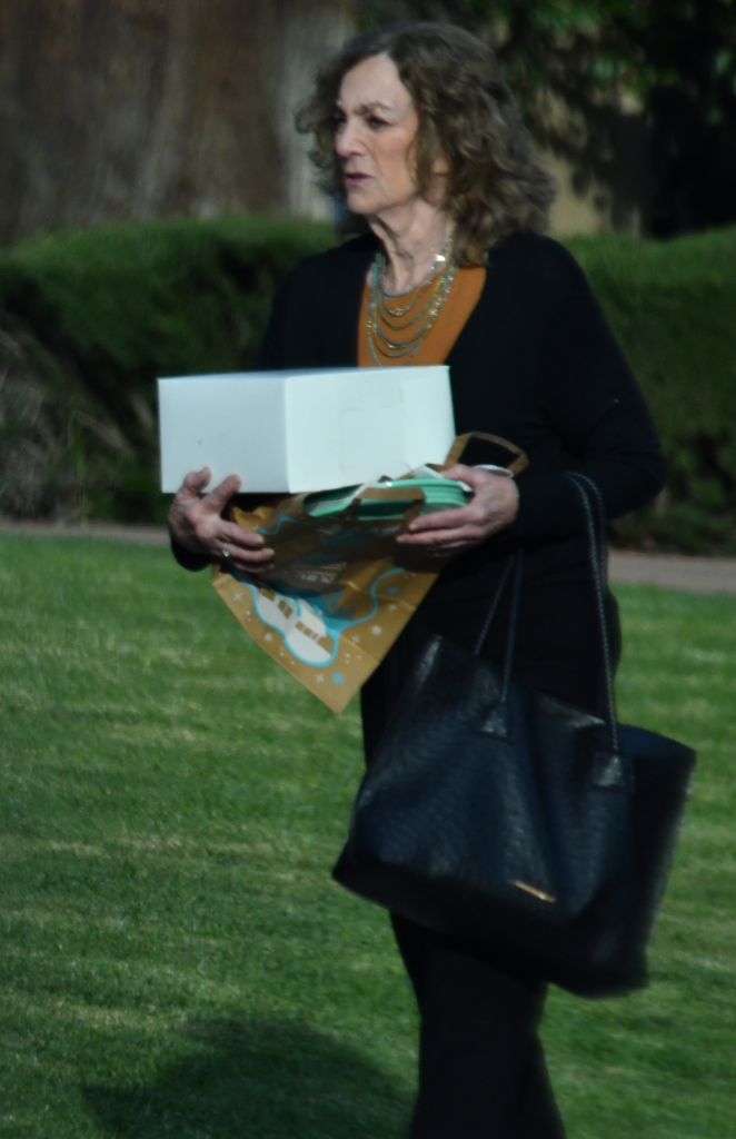 A woman holding a white box and a gift bag, along with a black handbag, walks across a lawn.